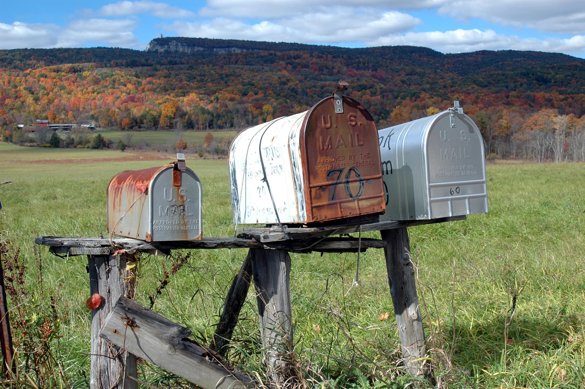 Four metal rural mailboxes standing in a row on a rough grey plank carried on splayed timber legs, one small and rust streaked, one pale and ribbed with writing along its side, one rust brown with U S MAIL and APPROVED BY THE POSTMASTER-GENERAL embossed on its door and a large hand painted 70 across the same door, and one silver with a small 60 written low on its door, all of them standing in long grass in front of a green field, with orange and gold autumn woodland rising behind it, two small farm buildings at the treeline and a long ridge closing the valley with a pale cliff face at its top