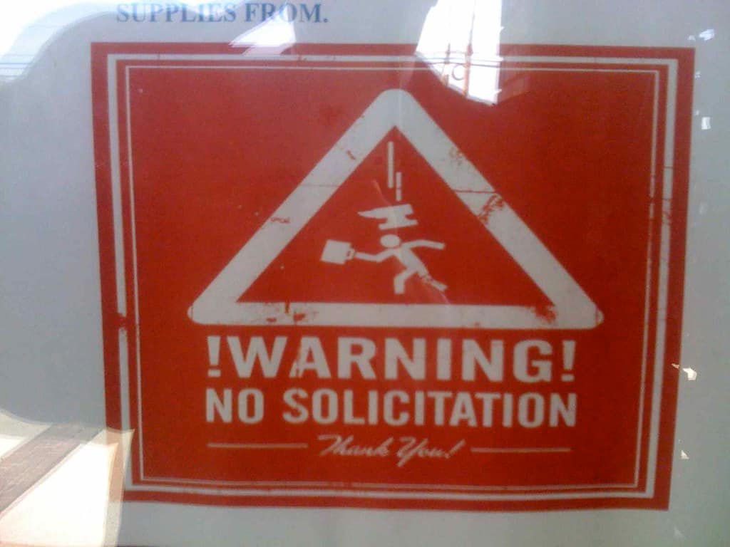 A weathered red metal sign filling the frame, scratched and faded, carrying a large white outlined triangle with a white pictogram inside it of a figure striding to the right with a case in one hand and its broad hat flying off above its head with a motion line over it, and beneath the triangle the words exclamation mark WARNING exclamation mark above the words NO SOLICITATION, both in white capitals, and below those, between two short rules, the words Thank You! in a white script hand, with a pale grey surface showing at each edge of the frame