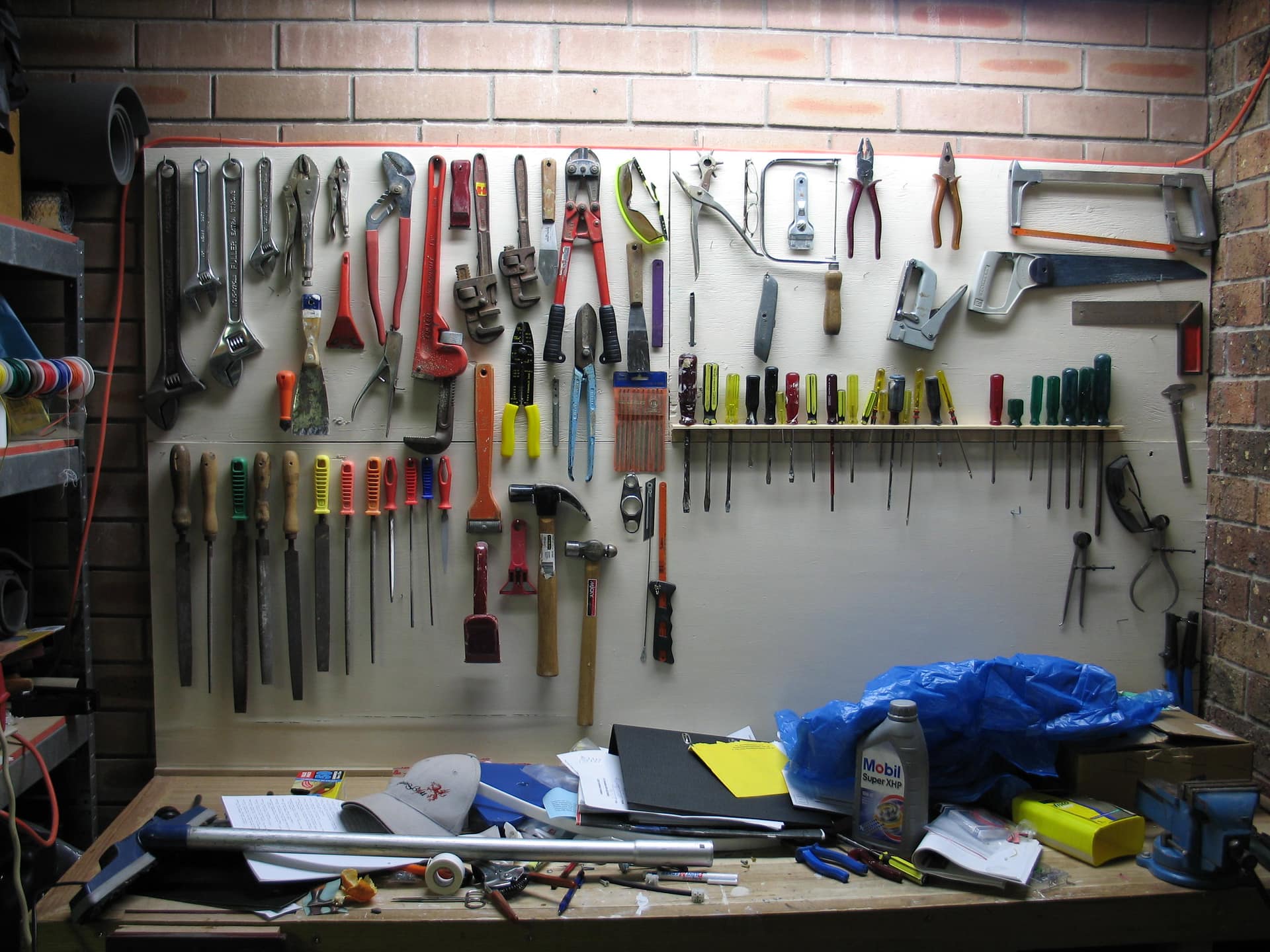 A workshop wall hung with hand tools in deliberate rows on pale painted boarding, adjustable spanners, pliers, a red pipe wrench and long handled bolt cutters along the top with a pair of yellow goggles among them, long files and rasps standing upright at the left, hammers and scrapers beside them, a shelf of screwdrivers with yellow, green, red and black handles ranked by size across the middle with chisels and punches hanging beneath it, two hand saws, a hacksaw frame and a large steel try square at the right against exposed brick with a second pair of goggles and two pairs of dividers below them, and a bench along the bottom of the frame carrying a blue plastic sheet, a labelled bottle of engine oil, a black folder and loose papers