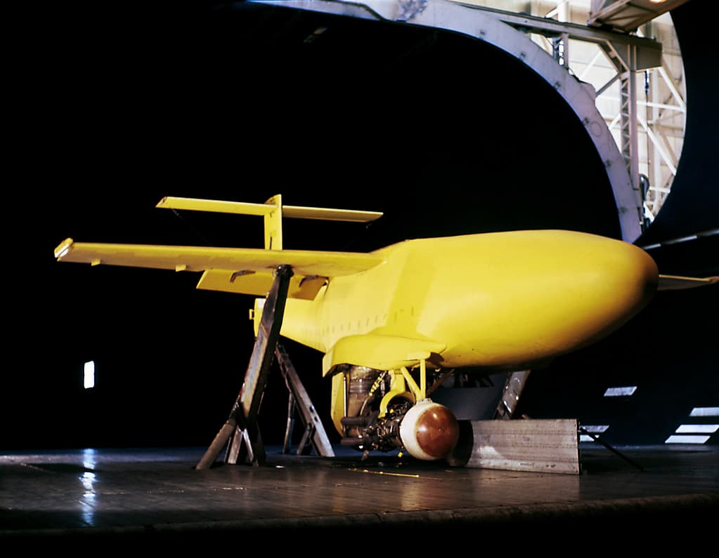 A yellow full scale aircraft model standing on trestles at the open mouth of a wind tunnel, its short wings held by a straight brace across them, exposed machinery under the fuselage and a rounded metal fairing resting on the deck beneath, with the dark curved throat of the tunnel behind it