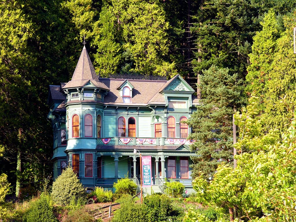 A green and teal painted Queen Anne Victorian with a pointed turret and tall arched windows in red painted surrounds glowing warm orange, a first-floor balcony hung with a row of red, white and blue fan bunting, a tall pink banner beside the entrance, standing against a bank of dark conifers