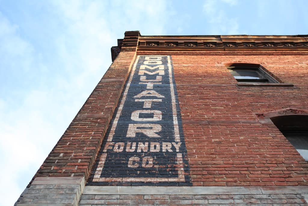 Looking steeply up a red brick wall at a tall painted sign in white capitals on a dark panel, its letters running vertically one above the next, the upper ones painted over each other so the name cannot be resolved and only the last of them standing clear and reading TOR, with FOUNDRY and CO in horizontal capitals below, a corbelled brick cornice along the top, a window in the brickwork to the right and pale blue sky along the left edge