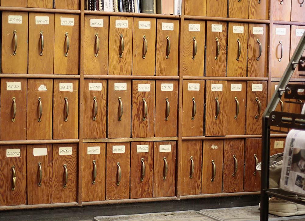 Three tiers of wooden card index drawers filling the frame, warm honey coloured varnished pine with long brass bail handles, each drawer carrying a small white card in a metal frame lettered by hand, the middle tier reading ranges of surnames such as ROC to RYZ and SAR to SGZ while the bottom tier reads DISTRICT twice and HANSARD four times with one card left blank, and a dark metal rack, a paler cabinet and a bundle of newspapers at the right hand edge