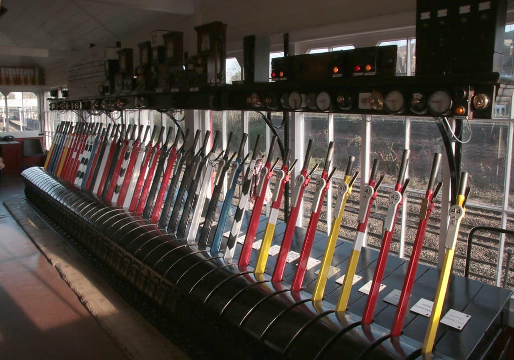 A long rank of tall painted signal levers in a railway signal box, receding away from the camera, most of them red with white, yellow, blue and black-and-white chequered levers standing among them, each with a curved catch handle at the top and a small numbered oval plate on its shaft, a row of brass instruments and dials mounted on the frame above them, a black lever bed and a plank floor below, and windows along the right showing track outside