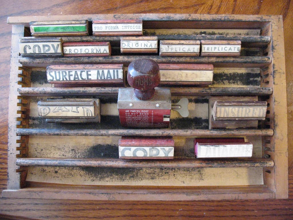 A worn wooden rack of rubber stamps photographed from above, the printed labels on their sides reading COPY, PROFORMA, ORIGINAL, DUPLICATE and TRIPLICATE along one row, SURFACE MAIL on a long stamp below them, PAID at the left and INSURED at the right of the row under that, and COPY again along the bottom, with a dark red wooden handled stamp standing in the middle whose red body is a rotary selector listing six settings, AIR PARCEL POST, DO NOT BEND, FIRST CLASS MAIL, FOR DEPOSIT ONLY, HAND STAMP ONLY and PARCEL POST, with a brass winged key at its side