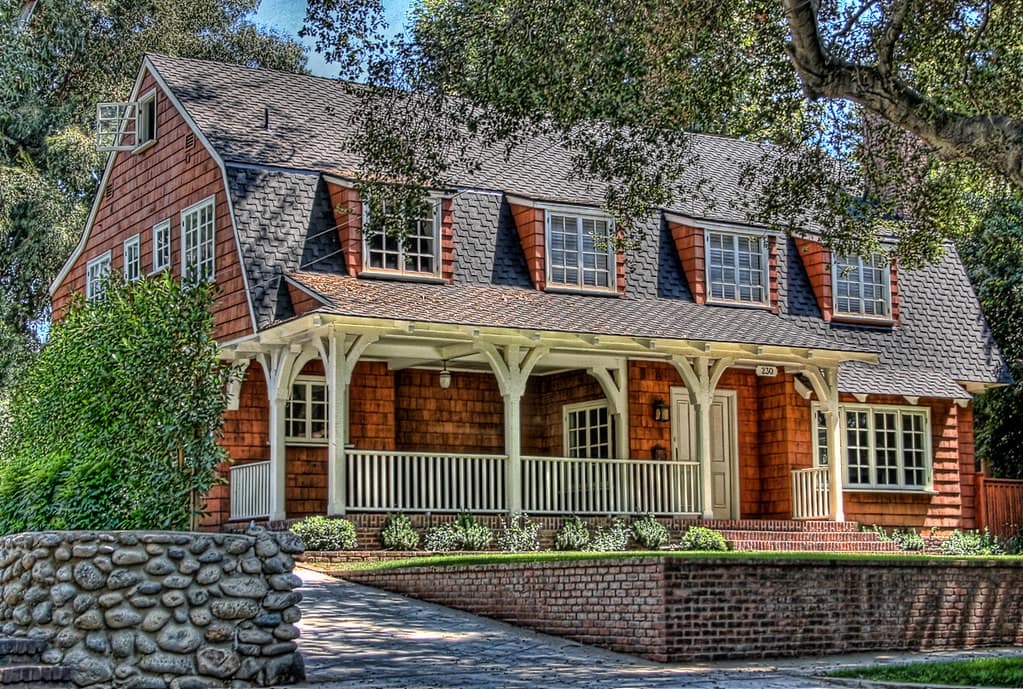 A cedar shingled house with a gambrel roof and a row of shingled dormers, a deep covered porch across the front carried on bracketed white posts with a lantern hanging under it, seen from the street over a river stone wall and a brick retaining wall, with mature trees crowding in on both sides