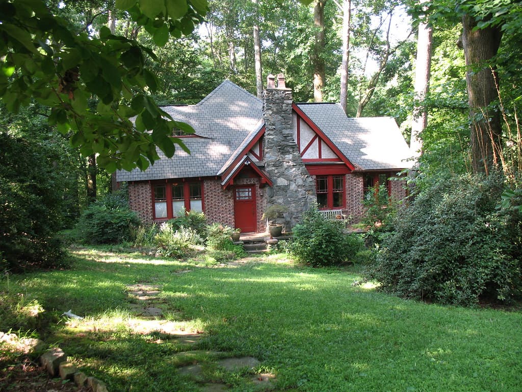 A brick and stone cottage with red-painted timbering, red window frames and a red front door, a wide stone chimney stack standing against the front of the house and rising past the roof, set in mature woodland behind a short mown lawn with deep shrub borders and a line of flat stepping stones leading toward the door